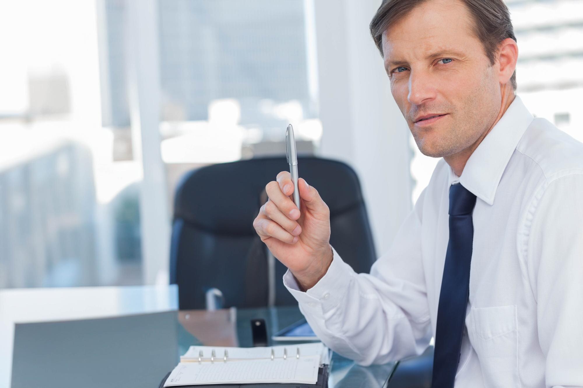 Businessman holding pen at office desk with binder and tablet and looking toward camera copy space